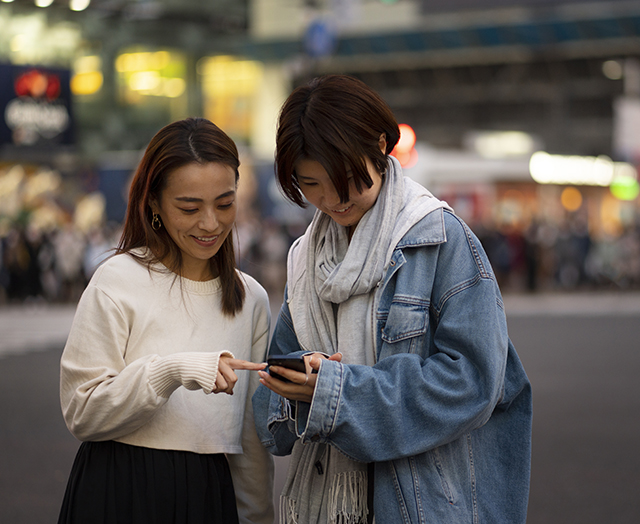華燈初上的都市熱鬧街鎮人行道背景下，兩位看起來氣質清新秀麗且面帶爽朗笑容的美妙妙齡粉領女子好友們結伴駐足，開心地將頭湊在一起查看一小塊共用手機螢幕上面的有趣內容的捕捉鏡頭