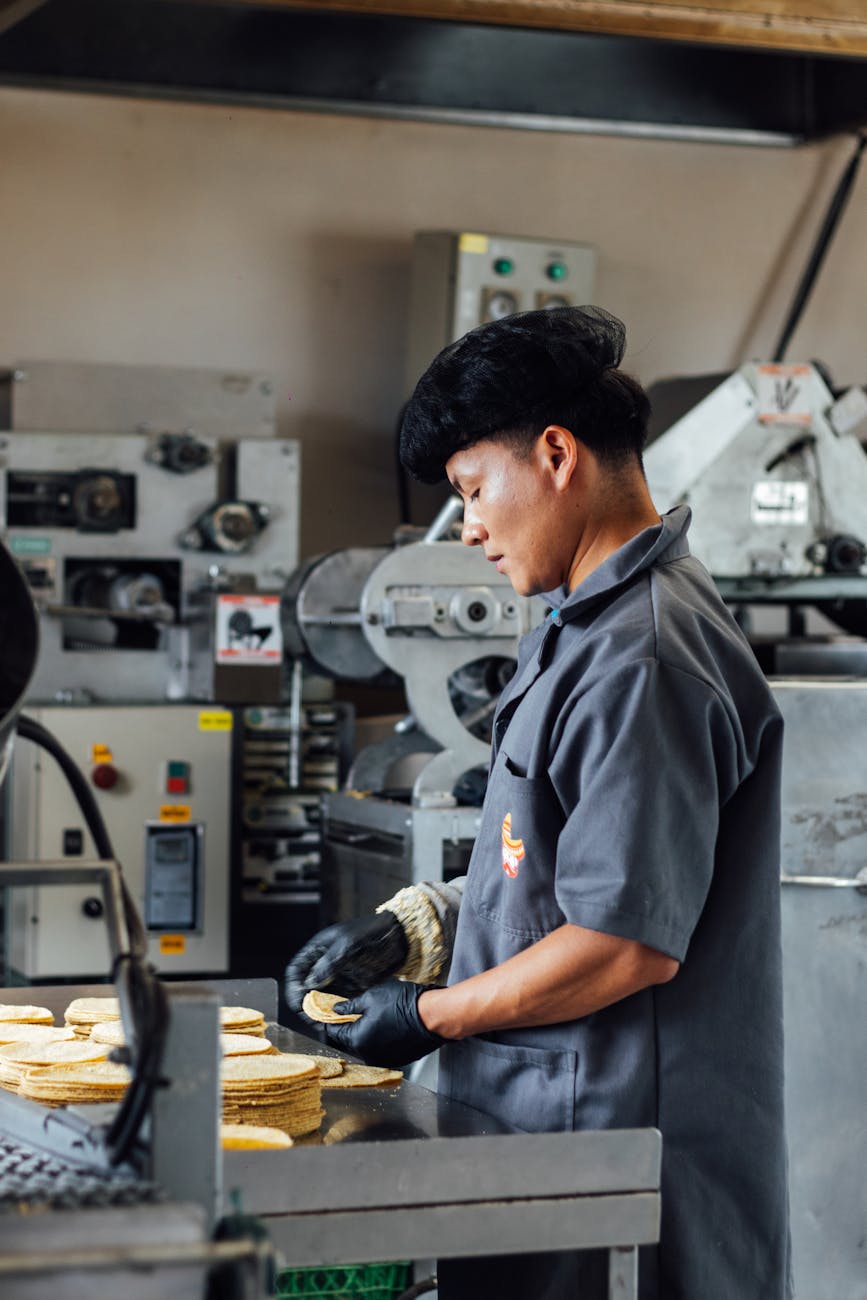 畫面正顯示出有位頭戴符合規範以阻擋落髮黑網罩、身兼穿灰色素色乾淨工作大衣這類工廠操作制服的資深中年男性從業員工職人的半身正面神情剪影照：圖中的他正面臨流水線上巨大繁複的自動機台組件和生產生鮮食材的各節點實施仔細而緊密的品管巡檢